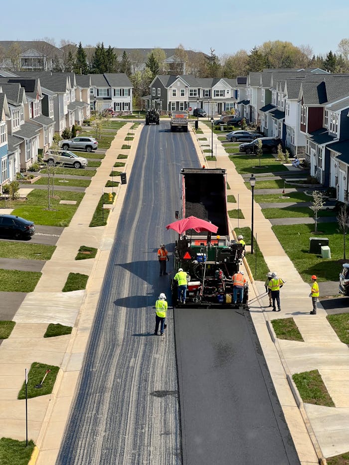 Workers repaving a street in a suburban residential neighborhood on a sunny day.